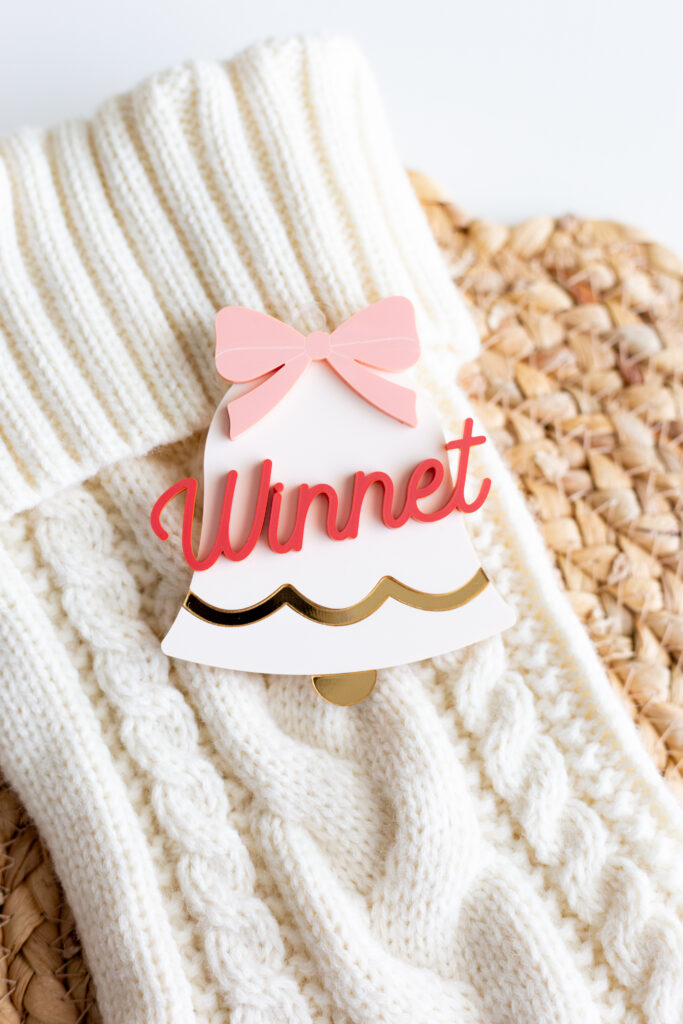 Laser cut Christmas ornament in the shape of a bell made out of acrylic and features a 3D name on top by Made of Embers